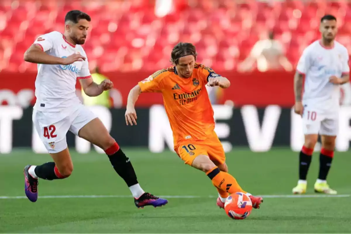 Two players compete for the ball in a match, one in a white uniform and the other in an orange uniform, while a third player watches from the background. Two players compete for the ball in a match, one in a white uniform and the other in an orange uniform, while a third player watches from the background.