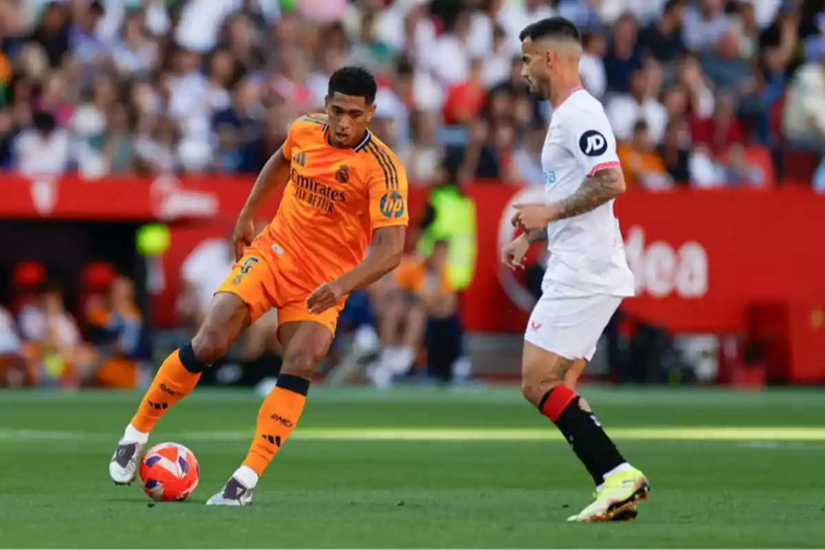 A player in an orange uniform controls the ball while an opponent in a white uniform watches in a stadium full of spectators.
