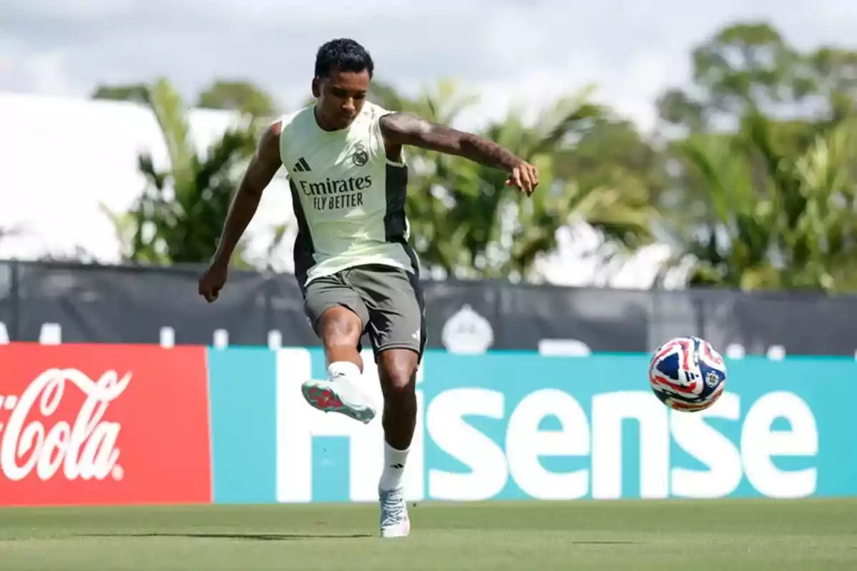 Soccer player training on a grass field while kicking a ball, with advertising in the background and palm trees.