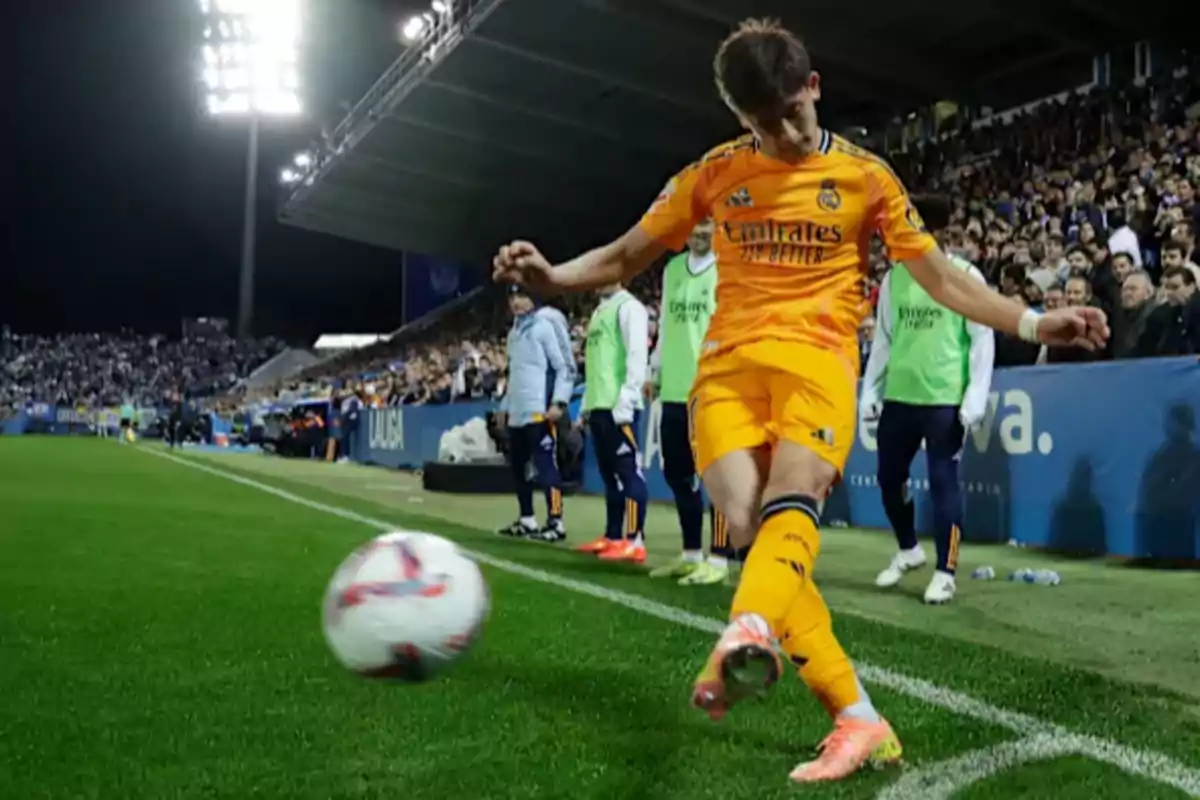 Soccer player in orange uniform kicking a ball near the touchline during a night match with spectators in the background.