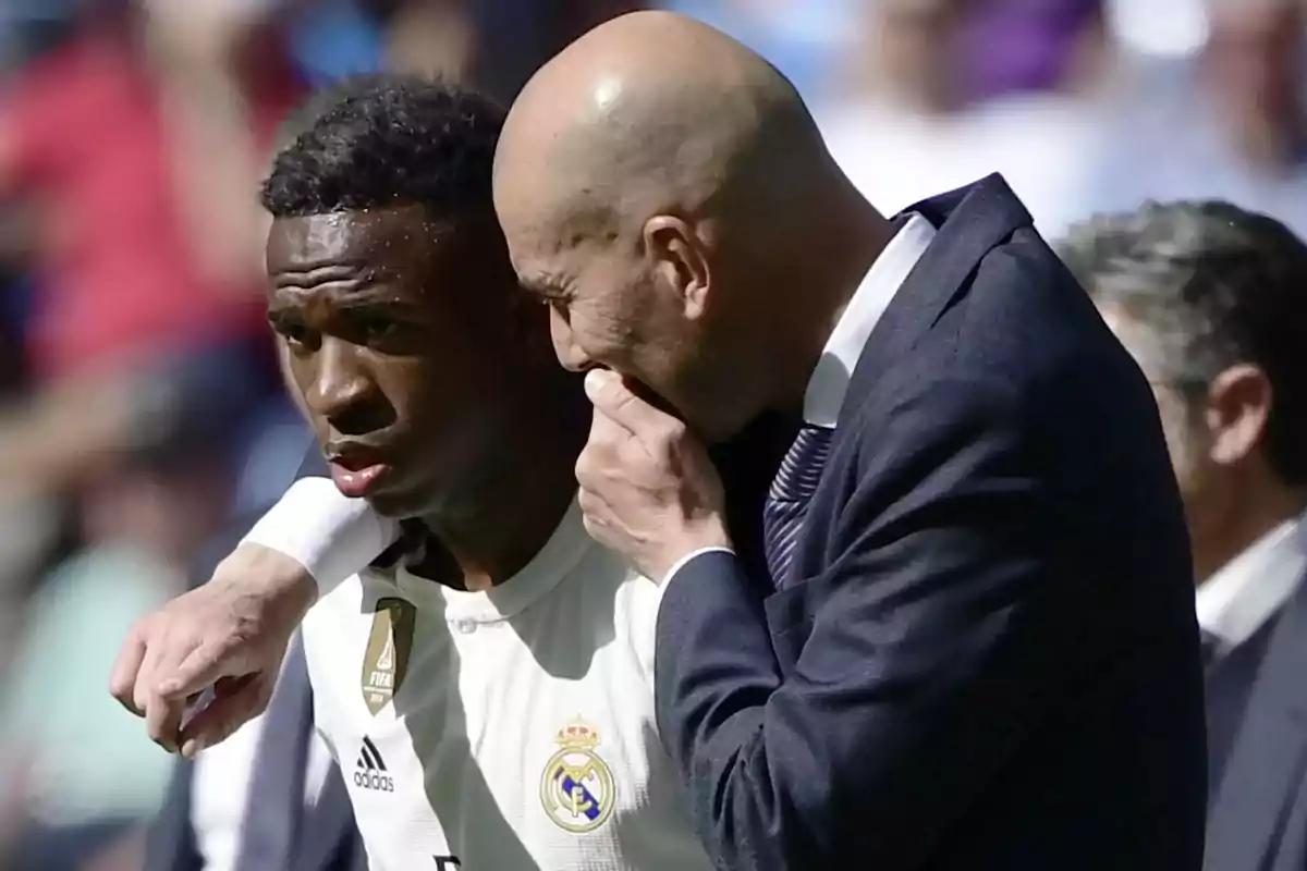 Un jugador de fútbol con el uniforme del Real Madrid escucha atentamente a un hombre calvo que le habla al oído en un campo de fútbol.