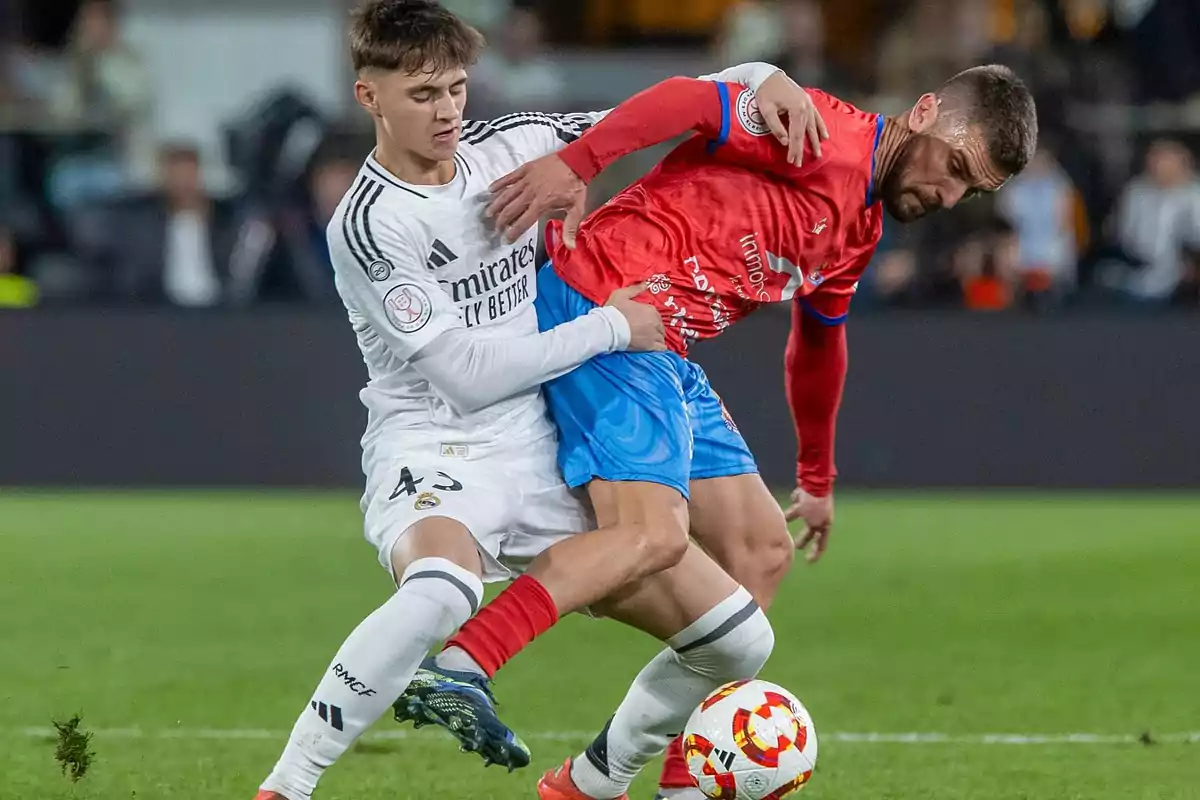 Two soccer players compete for the ball in a game, one wearing a white uniform and the other wearing a red and blue uniform. Two soccer players compete for the ball in a game, one wearing a white uniform and the other wearing a red and blue uniform.