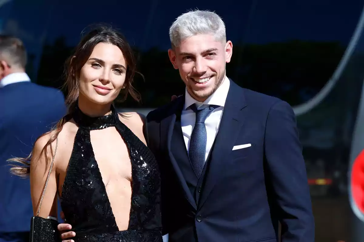 Una pareja vestida elegantemente sonríe mientras posa para una foto en un evento al aire libre.