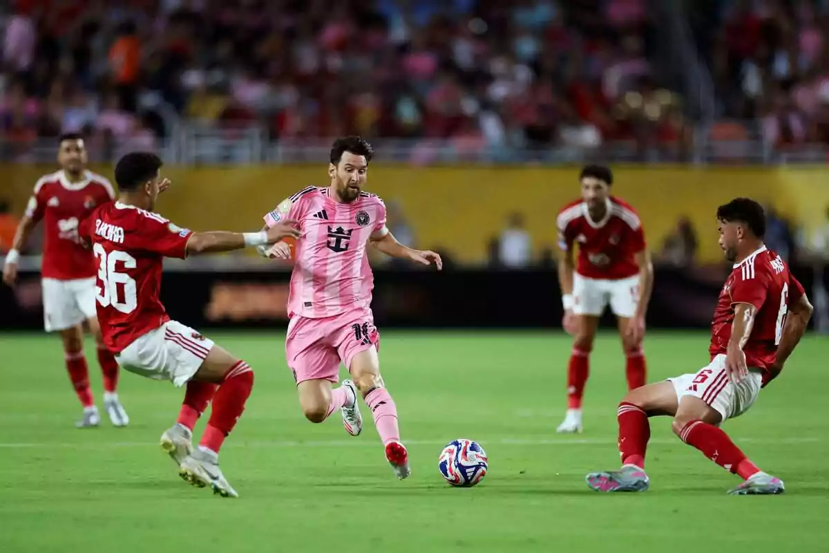 Un jugador con uniforme rosa avanza con el balón mientras es rodeado por tres rivales con uniforme rojo en un partido de fútbol. Un jugador con uniforme rosa avanza con el balón mientras es rodeado por tres rivales con uniforme rojo en un partido de fútbol.