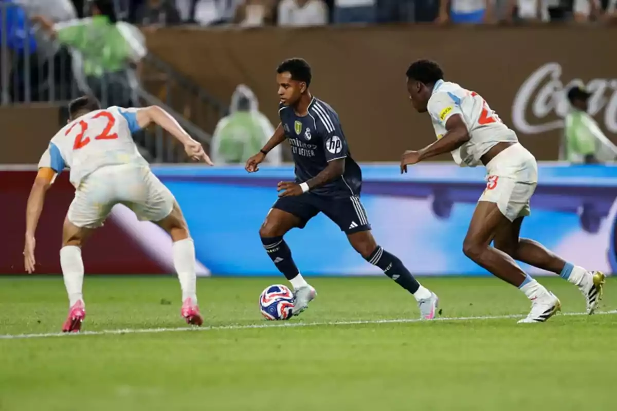 Un jugador con uniforme oscuro controla el balón mientras dos rivales con uniforme blanco intentan detenerlo en un partido de fútbol. Un jugador con uniforme oscuro controla el balón mientras dos rivales con uniforme blanco intentan detenerlo en un partido de fútbol.