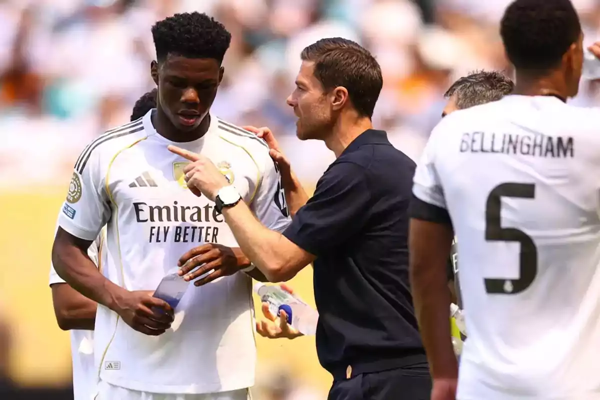 Entrenador dando indicaciones a un jugador del Real Madrid durante un partido mientras otro jugador bebe agua