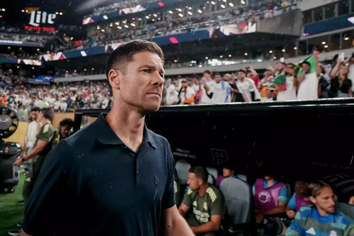 Hombre con camisa azul oscuro caminando frente a un banquillo de fútbol en un estadio lleno de aficionados Hombre con camisa azul oscuro caminando frente a un banquillo de fútbol en un estadio lleno de aficionados