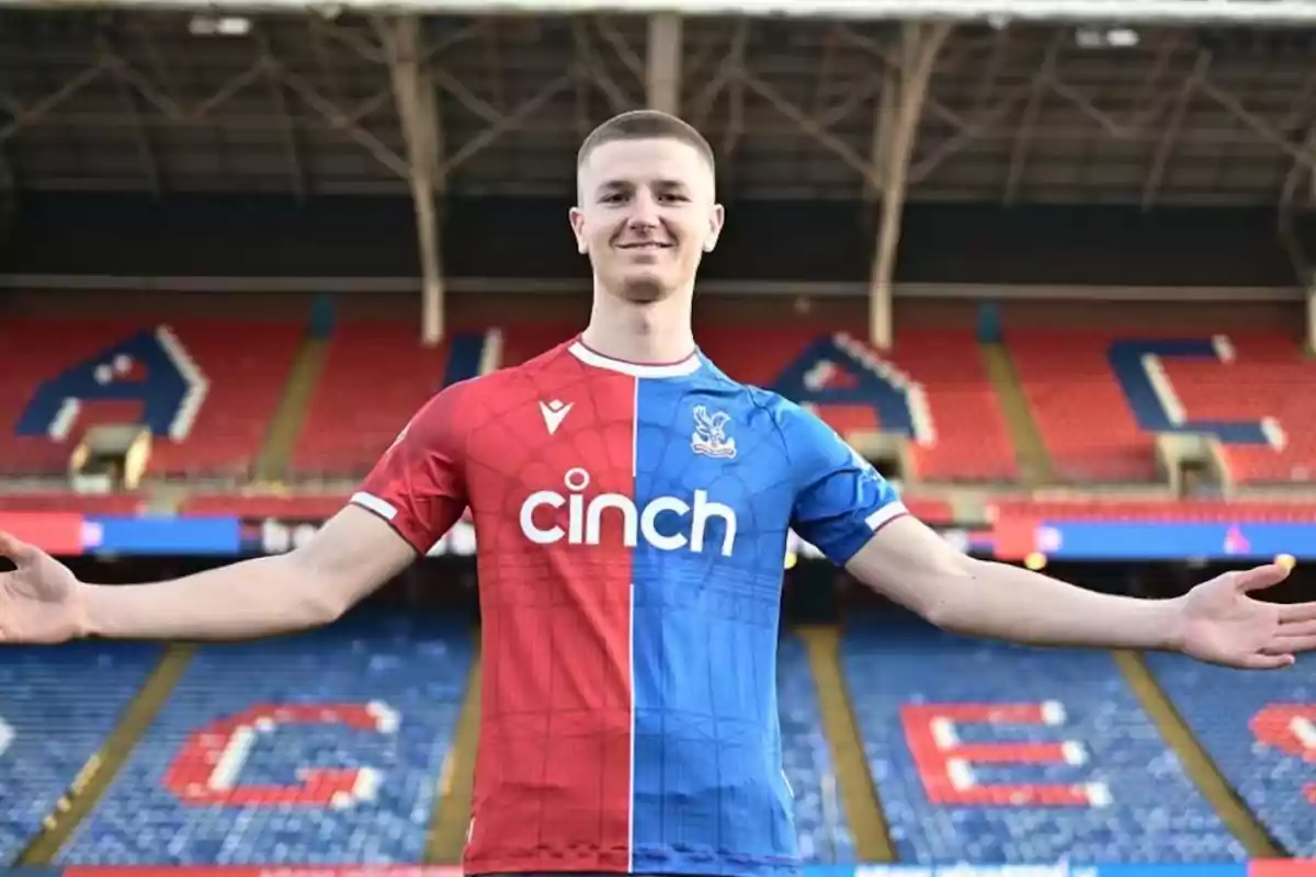 Jugador de fútbol con los brazos extendidos usando el uniforme rojo y azul del Crystal Palace en un estadio vacío Jugador de fútbol con los brazos extendidos usando el uniforme rojo y azul del Crystal Palace en un estadio vacío