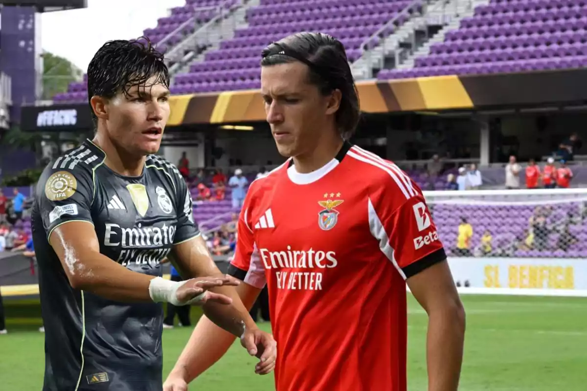 Dos futbolistas en el campo, uno con uniforme negro del Real Madrid y otro con uniforme rojo del Benfica, en un estadio con gradas moradas de fondo Dos futbolistas en el campo, uno con uniforme negro del Real Madrid y otro con uniforme rojo del Benfica, en un estadio con gradas moradas de fondo