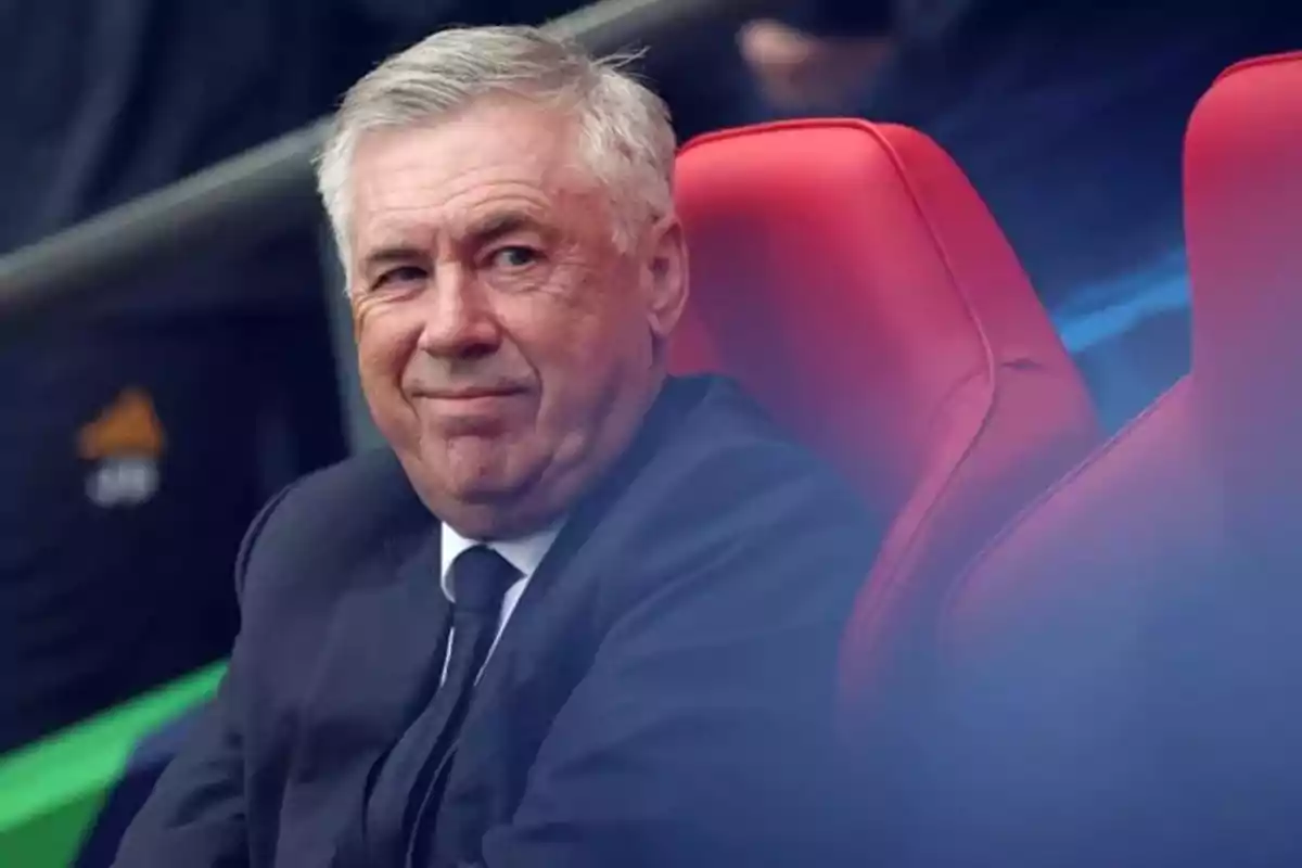 Gray-haired man in a dark suit sitting on a red chair, looking to the side and slightly smiling