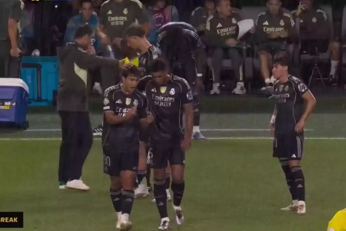 Real Madrid players in black uniforms talk and get ready on the field under the rain while the coaching staff and other players watch from the bench. Real Madrid players in black uniforms talk and get ready on the field under the rain while the coaching staff and other players watch from the bench.