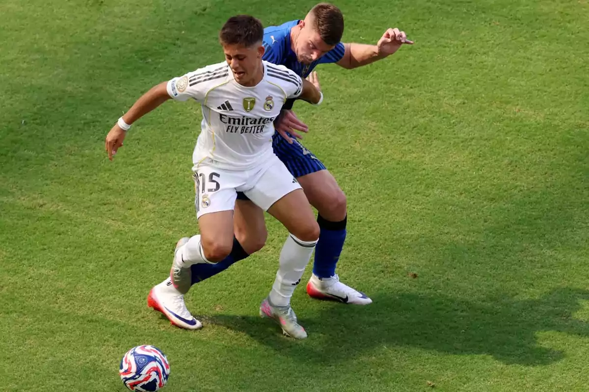 Two players compete for the ball in a match on grass, one wearing a white uniform and the other wearing a blue uniform. Two players compete for the ball in a match on grass, one wearing a white uniform and the other wearing a blue uniform.