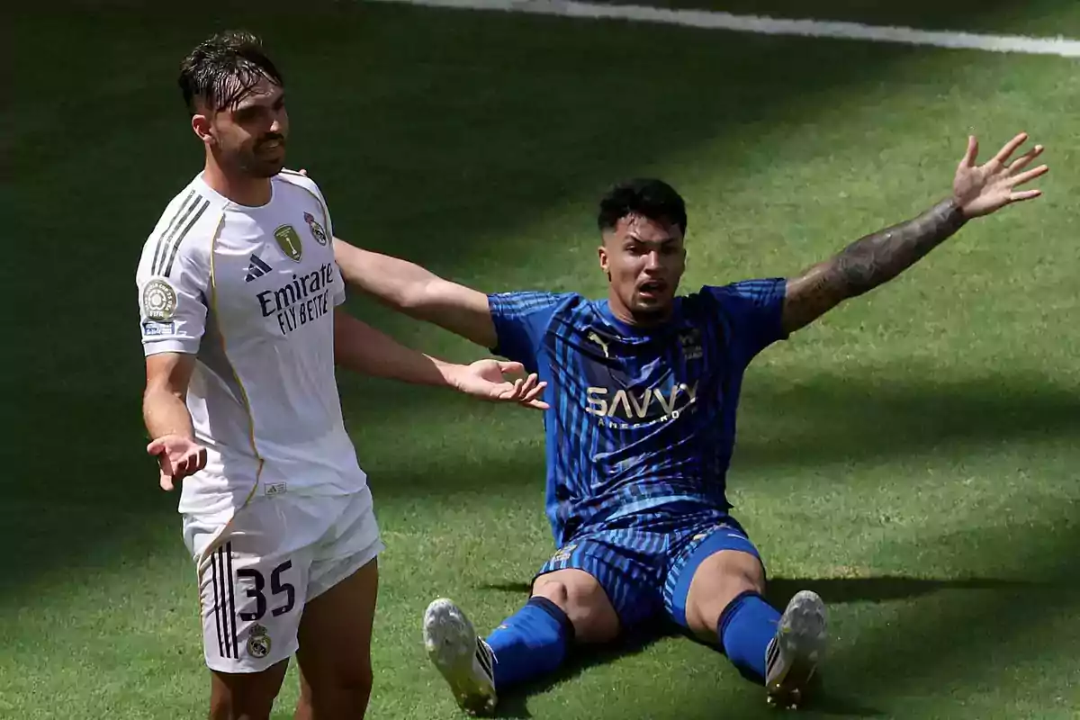 A player in a white uniform standing and another in a blue uniform sitting on the grass with his arms raised, both showing gestures of protest during a match. A player in a white uniform standing and another in a blue uniform sitting on the grass with his arms raised, both showing gestures of protest during a match.