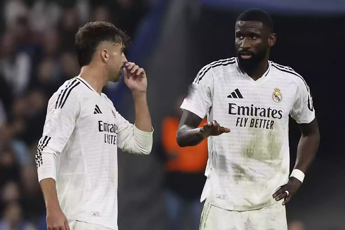 Dos futbolistas del Real Madrid conversan en el campo durante un partido, ambos llevan uniforme blanco y parecen concentrados en la estrategia. Dos futbolistas del Real Madrid conversan en el campo durante un partido, ambos llevan uniforme blanco y parecen concentrados en la estrategia.