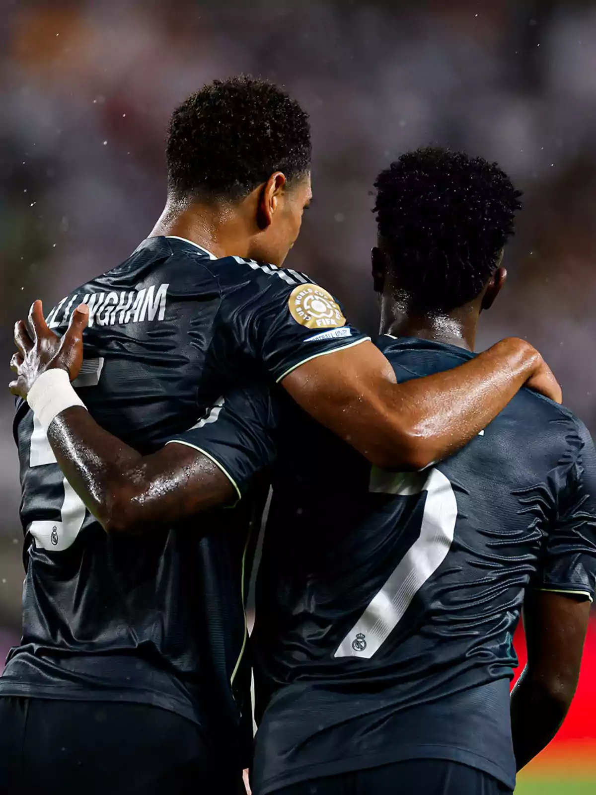 Two Real Madrid players are hugging in celebration during a match, both wearing dark uniforms and seen from behind.