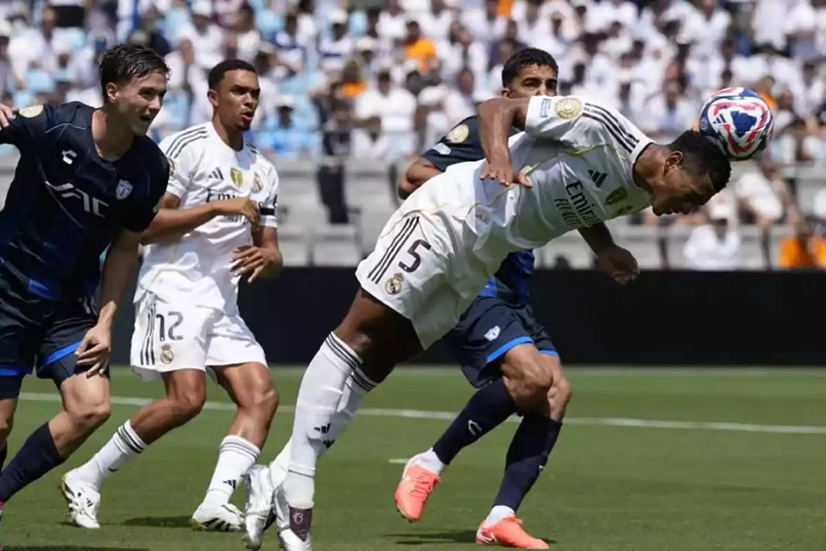 Futbolista con uniforme blanco remata el balón de cabeza durante un partido mientras varios jugadores lo rodean en el campo Futbolista con uniforme blanco remata el balón de cabeza durante un partido mientras varios jugadores lo rodean en el campo
