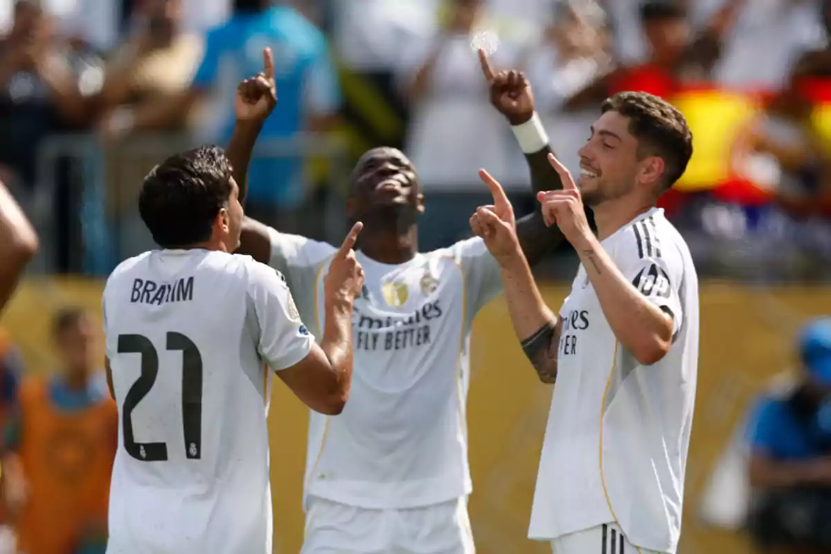 Tres jugadores del Real Madrid celebran un gol señalando al cielo y sonriendo en el campo de fútbol