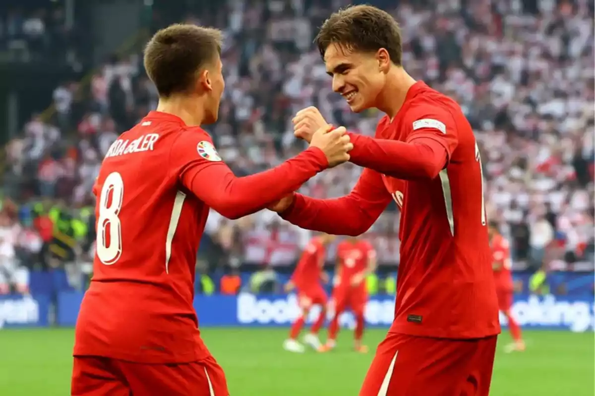 Two players in red uniforms celebrate together on the field during a match, surrounded by a stadium full of fans. Two players in red uniforms celebrate together on the field during a match, surrounded by a stadium full of fans.