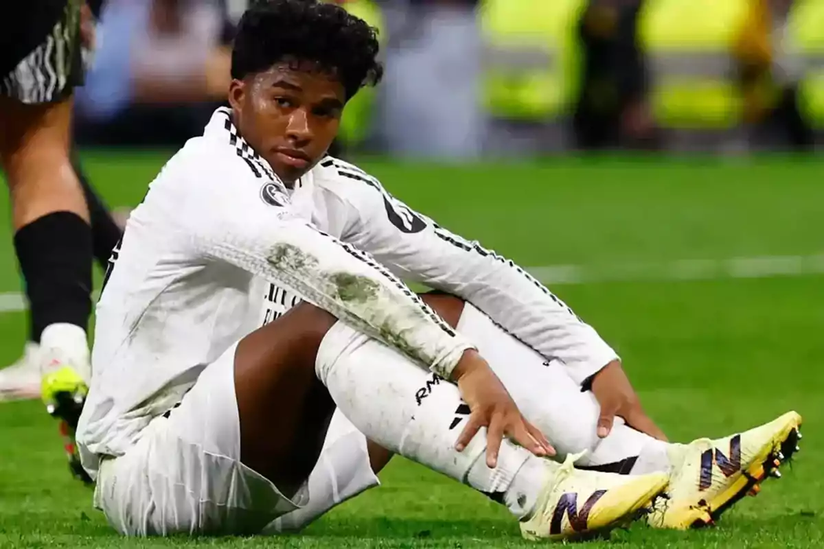 Futbolista sentado en el césped con uniforme blanco y manchas de tierra en la ropa, mostrando una expresión seria durante un partido. Futbolista sentado en el césped con uniforme blanco y manchas de tierra en la ropa, mostrando una expresión seria durante un partido.