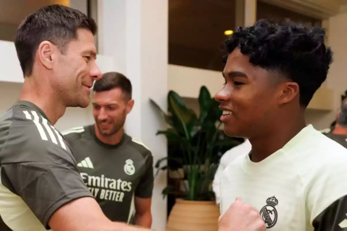 Two people wearing Real Madrid training gear greet each other with smiles inside a building while another person watches in the background. Two people wearing Real Madrid training gear greet each other with smiles inside a building while another person watches in the background.