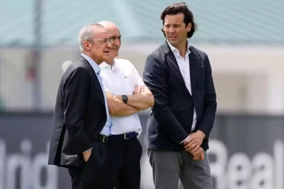 Three men standing and talking on a football training field, two of them wearing jackets and one wearing a white jersey.