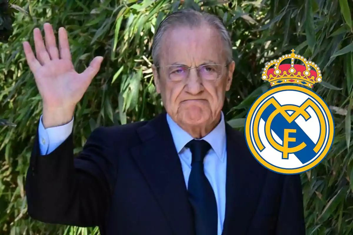 Hombre mayor con gafas y traje oscuro levantando la mano junto al escudo del Real Madrid frente a un fondo de plantas verdes