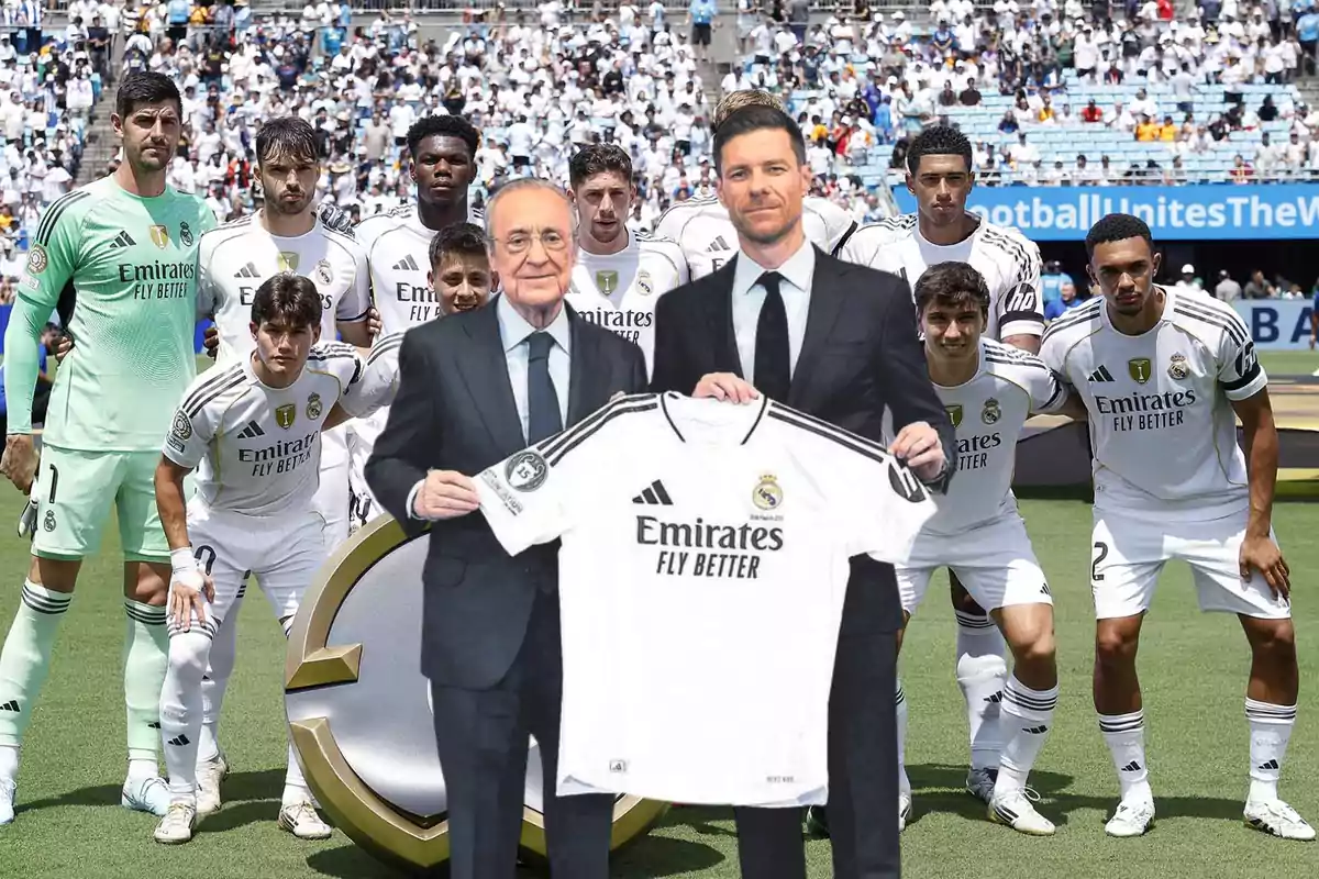 Dos hombres con traje sostienen una camiseta del Real Madrid frente a un grupo de jugadores del equipo en el campo de fútbol con el estadio lleno de aficionados al fondo