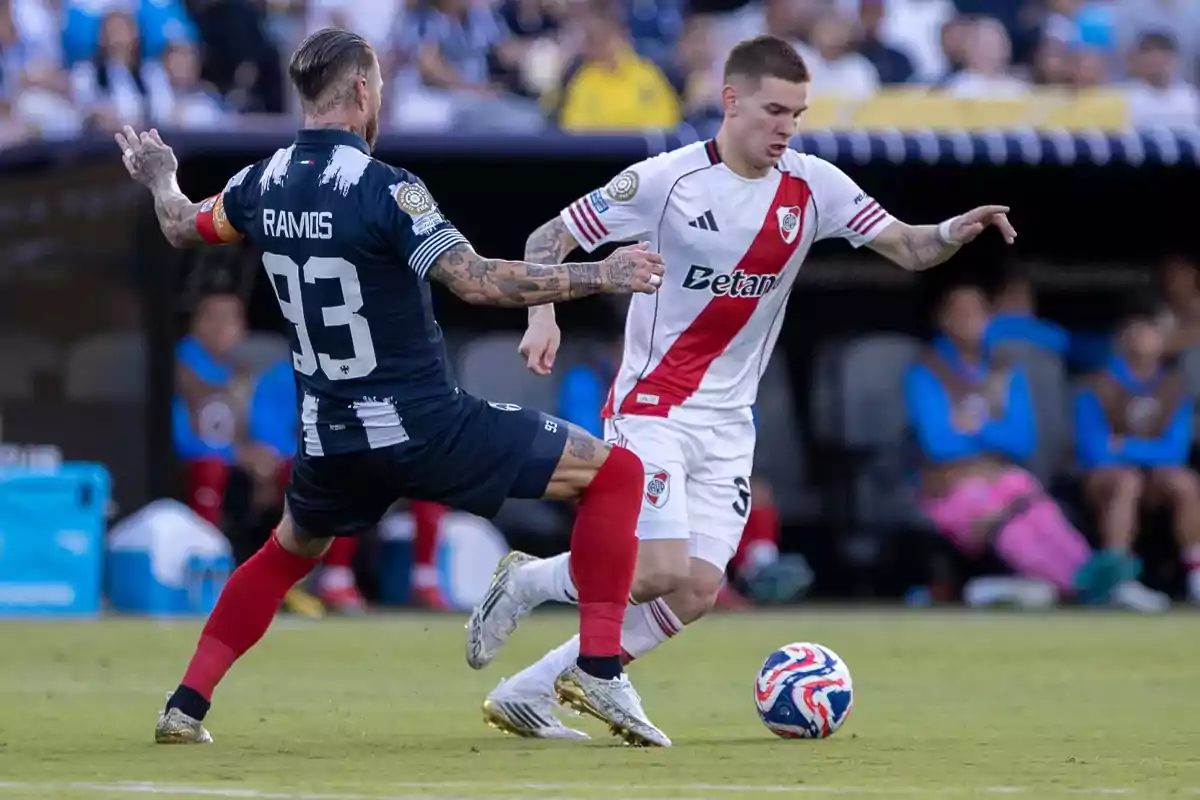 Two players compete for the ball during a match, one wearing a blue uniform and the other wearing a white uniform with a red stripe. Two players compete for the ball during a match, one wearing a blue uniform and the other wearing a white uniform with a red stripe.