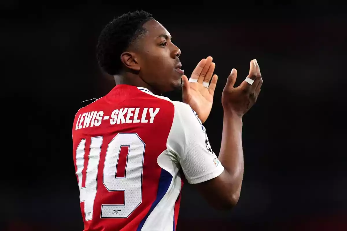 Soccer player in Arsenal’s red and white uniform applauding during a match, with the name Lewis-Skelly and the number 49 on the jersey. Soccer player in Arsenal’s red and white uniform applauding during a match, with the name Lewis-Skelly and the number 49 on the jersey.