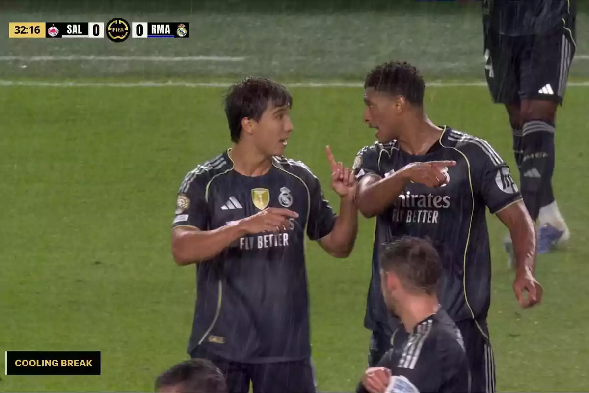 Two Real Madrid players talk and gesture in the rain during a hydration break in a soccer match, both wearing black uniforms, with the scoreboard showing zero in the background.