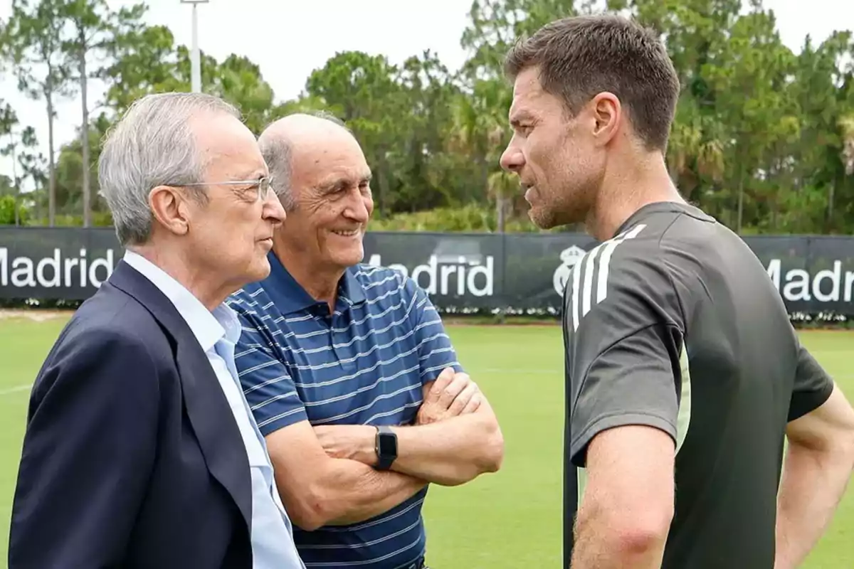 Tres hombres conversan en un campo de entrenamiento de fútbol con árboles y un cartel de Madrid al fondo Tres hombres conversan en un campo de entrenamiento de fútbol con árboles y un cartel de Madrid al fondo