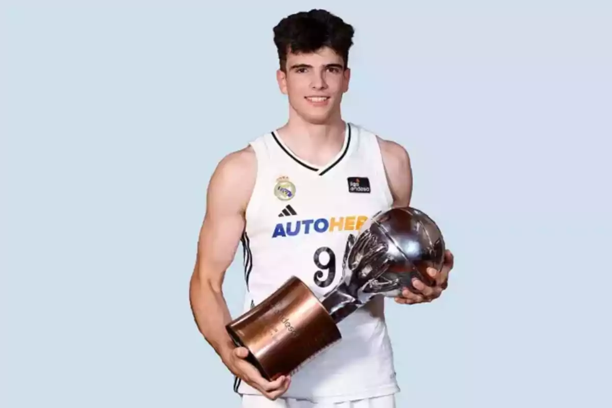 Basketball player in a white uniform holding a large silver trophy against a light blue background Basketball player in a white uniform holding a large silver trophy against a light blue background