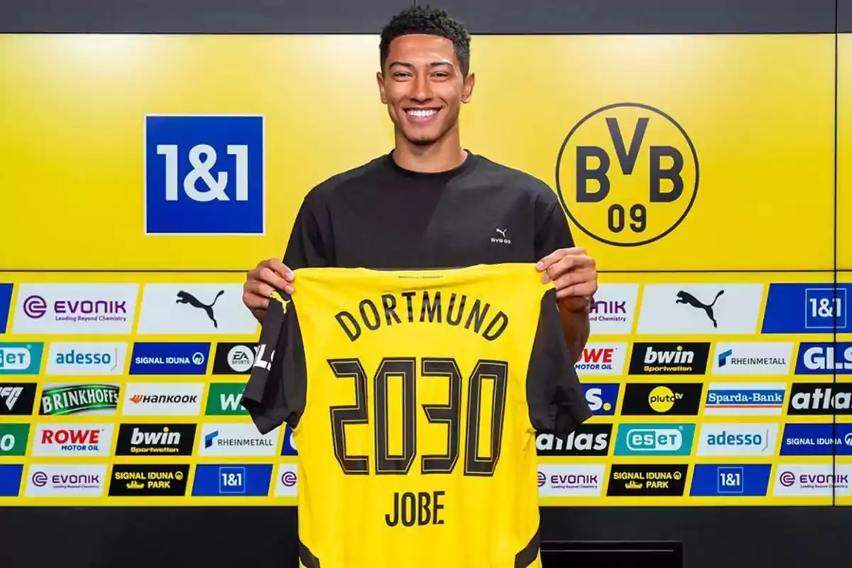 Jugador sonriente sostiene una camiseta amarilla del Borussia Dortmund con el nombre Jobe y el número 2030 en una sala de prensa con logotipos de patrocinadores al fondo