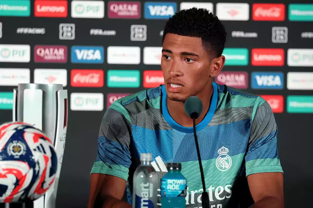 Futbolista del Real Madrid dando una conferencia de prensa con un micrófono frente a él, una botella de agua, una bebida deportiva, un trofeo y un balón en primer plano, con un fondo de patrocinadores.