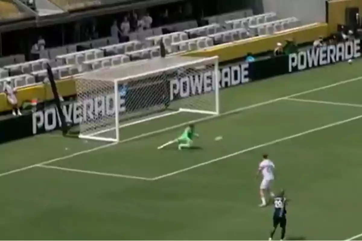 Portero lanzándose para atajar un balón durante un partido de fútbol con la portería de fondo y jugadores cerca del área Portero lanzándose para atajar un balón durante un partido de fútbol con la portería de fondo y jugadores cerca del área