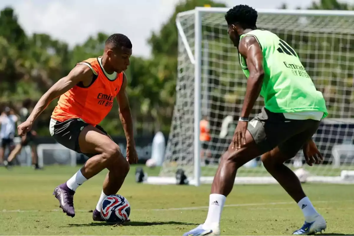 Two players are training on a soccer field; one wearing an orange bib controls the ball while the other, in a green bib, marks him in front of a goal. Two players are training on a soccer field; one wearing an orange bib controls the ball while the other, in a green bib, marks him in front of a goal.