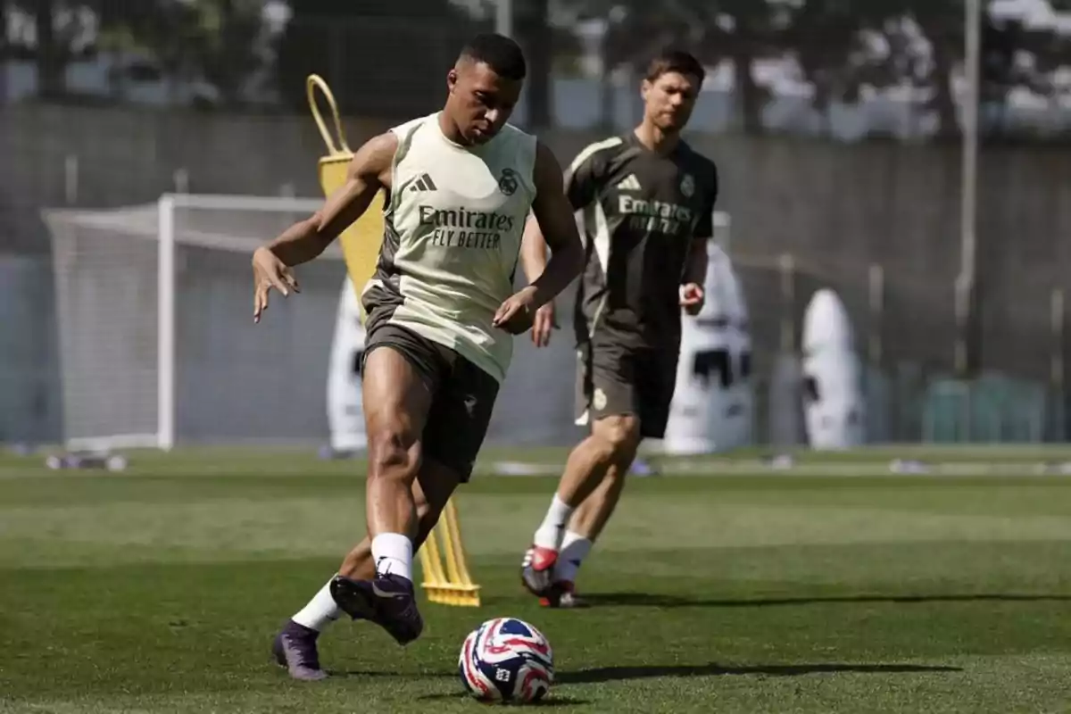 Futbolista entrenando en un campo de fútbol con un balón mientras otro jugador lo observa al fondo Futbolista entrenando en un campo de fútbol con un balón mientras otro jugador lo observa al fondo
