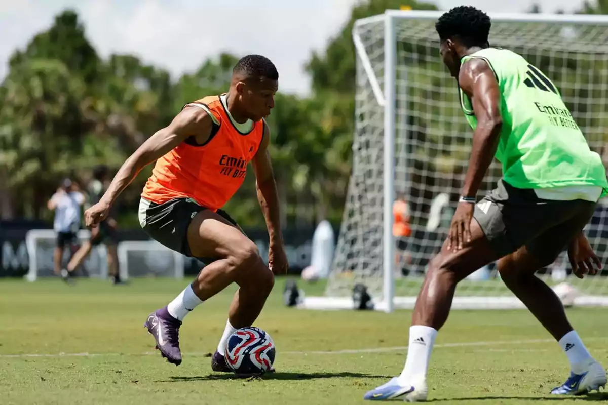 Two players are training on a grass field; one wearing an orange bib controls the ball while the other, in a green bib, marks him in front of a goal.