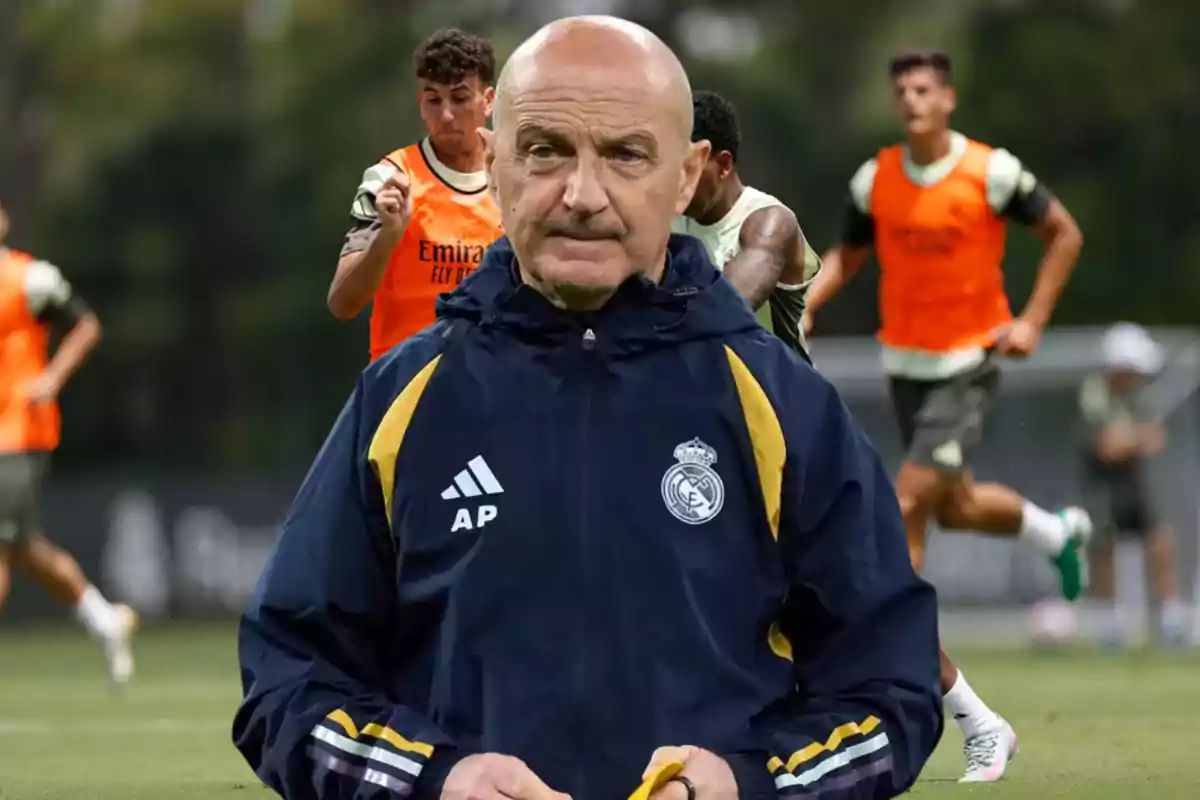 Real Madrid coach wearing a navy blue jacket with players training in the background on a soccer field