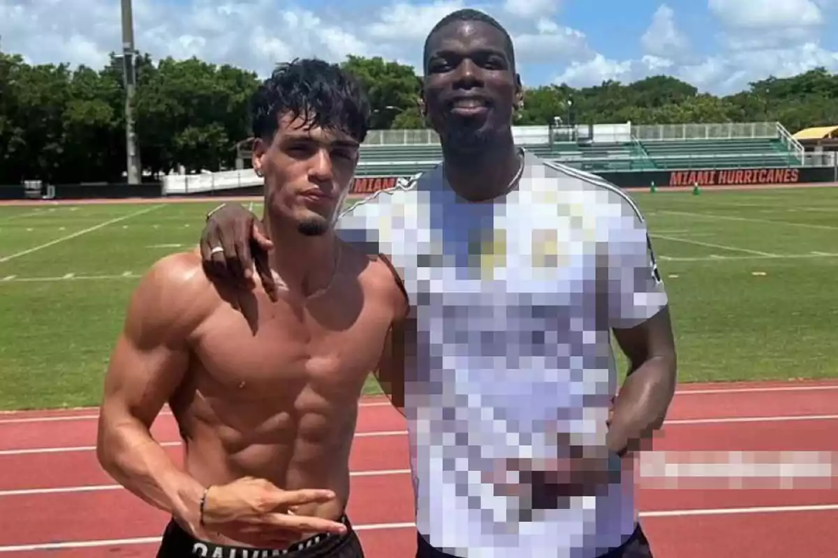 Dos hombres posan juntos en una pista de atletismo uno de ellos está sin camiseta mostrando su físico musculoso mientras el otro lleva puesta una camiseta deportiva y sonríe