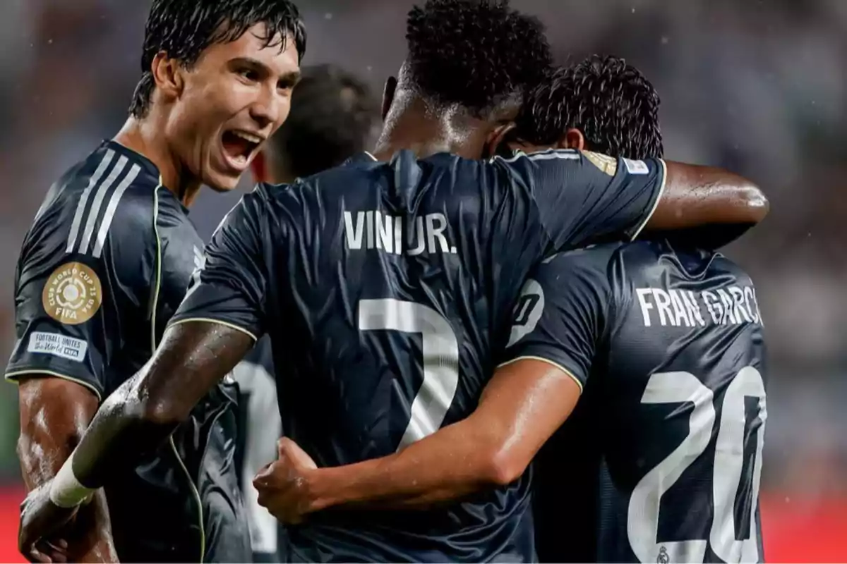 Tres futbolistas con uniforme oscuro celebran un gol bajo la lluvia en un partido, uno de ellos grita emocionado mientras los otros dos se abrazan Tres futbolistas con uniforme oscuro celebran un gol bajo la lluvia en un partido, uno de ellos grita emocionado mientras los otros dos se abrazan