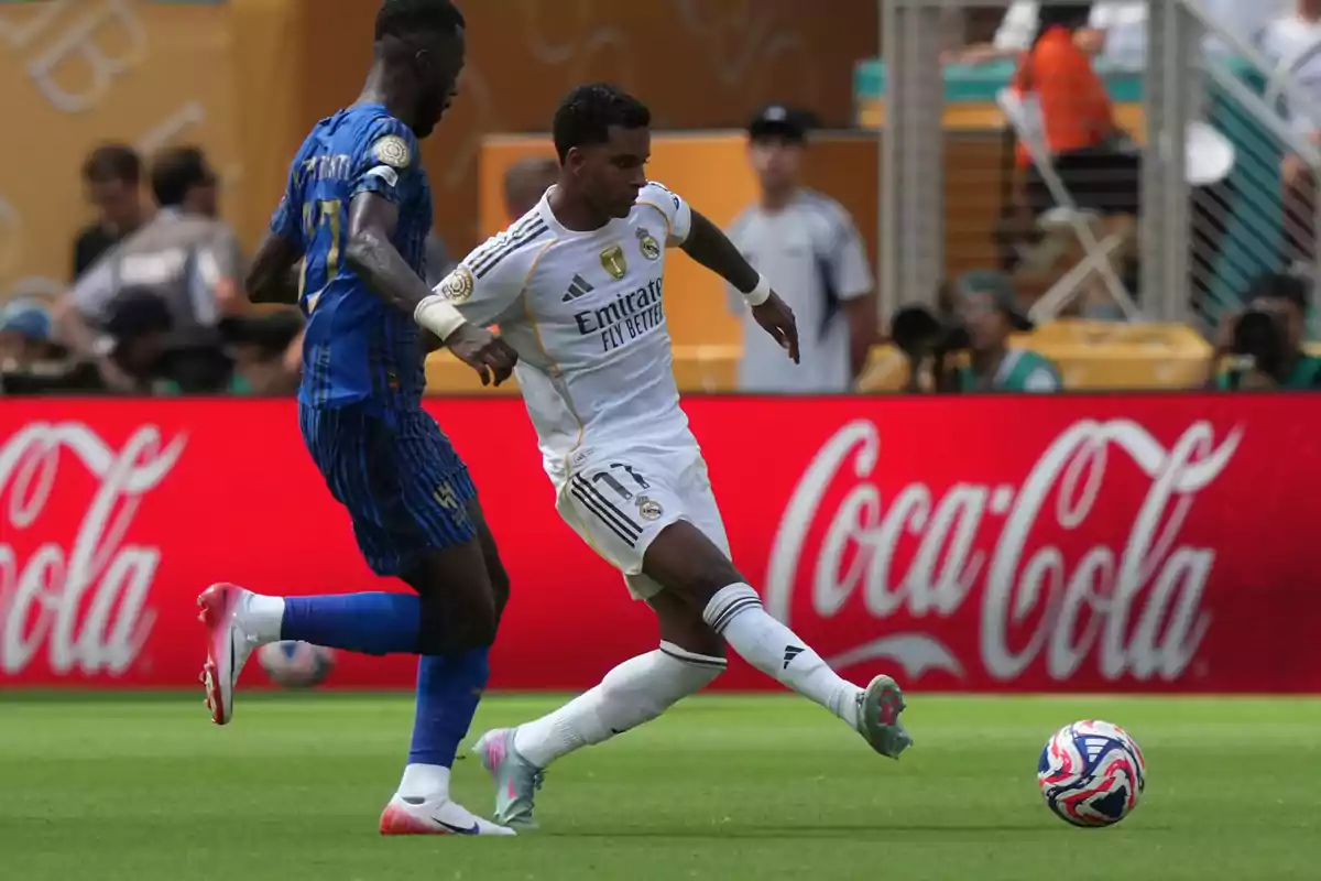 Dos futbolistas disputan el balón durante un partido en un estadio, uno con uniforme blanco y otro con uniforme azul.