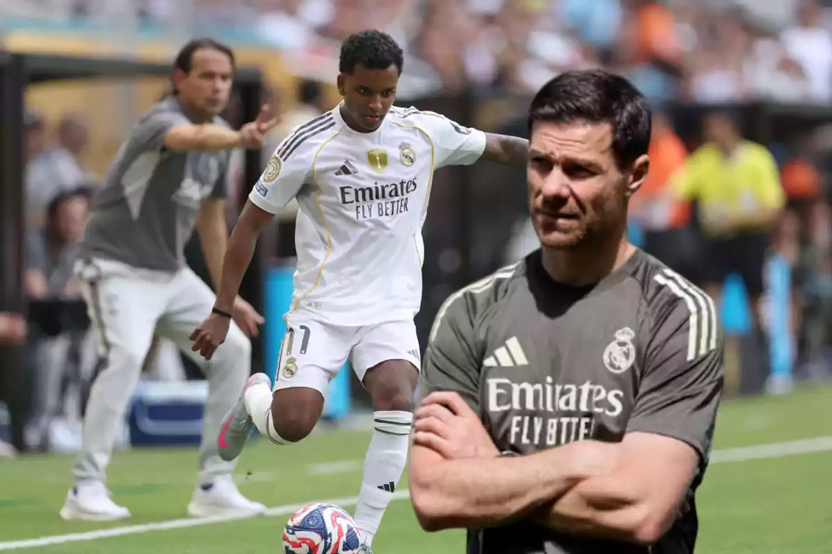 Un jugador del Real Madrid controla el balón en el campo mientras un entrenador observa y da indicaciones en el fondo y otro entrenador aparece en primer plano con los brazos cruzados