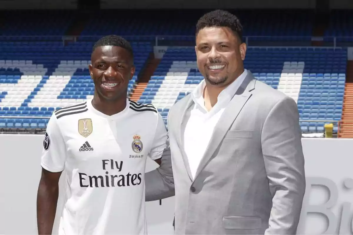 Two people are smiling in a soccer stadium, one is wearing a Real Madrid uniform and the other is dressed in a light gray suit.