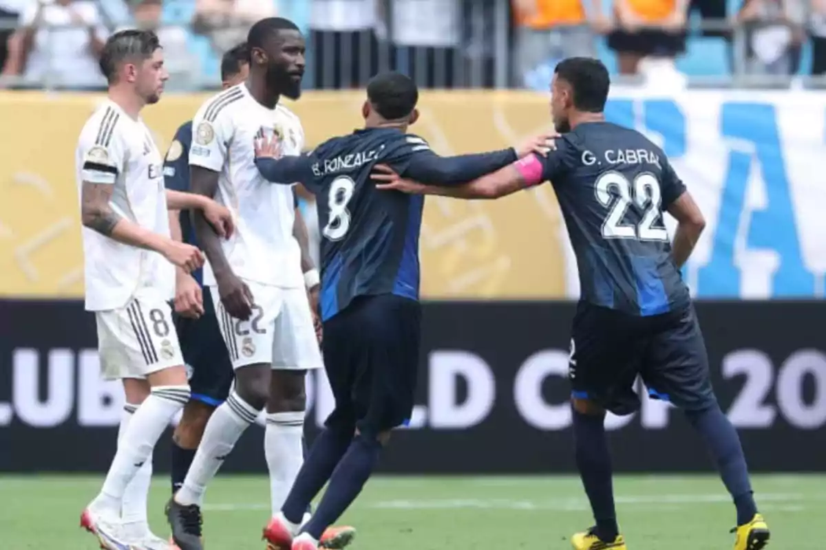 Four players argue on the field during a soccer match, two wearing white uniforms and two wearing dark blue uniforms.