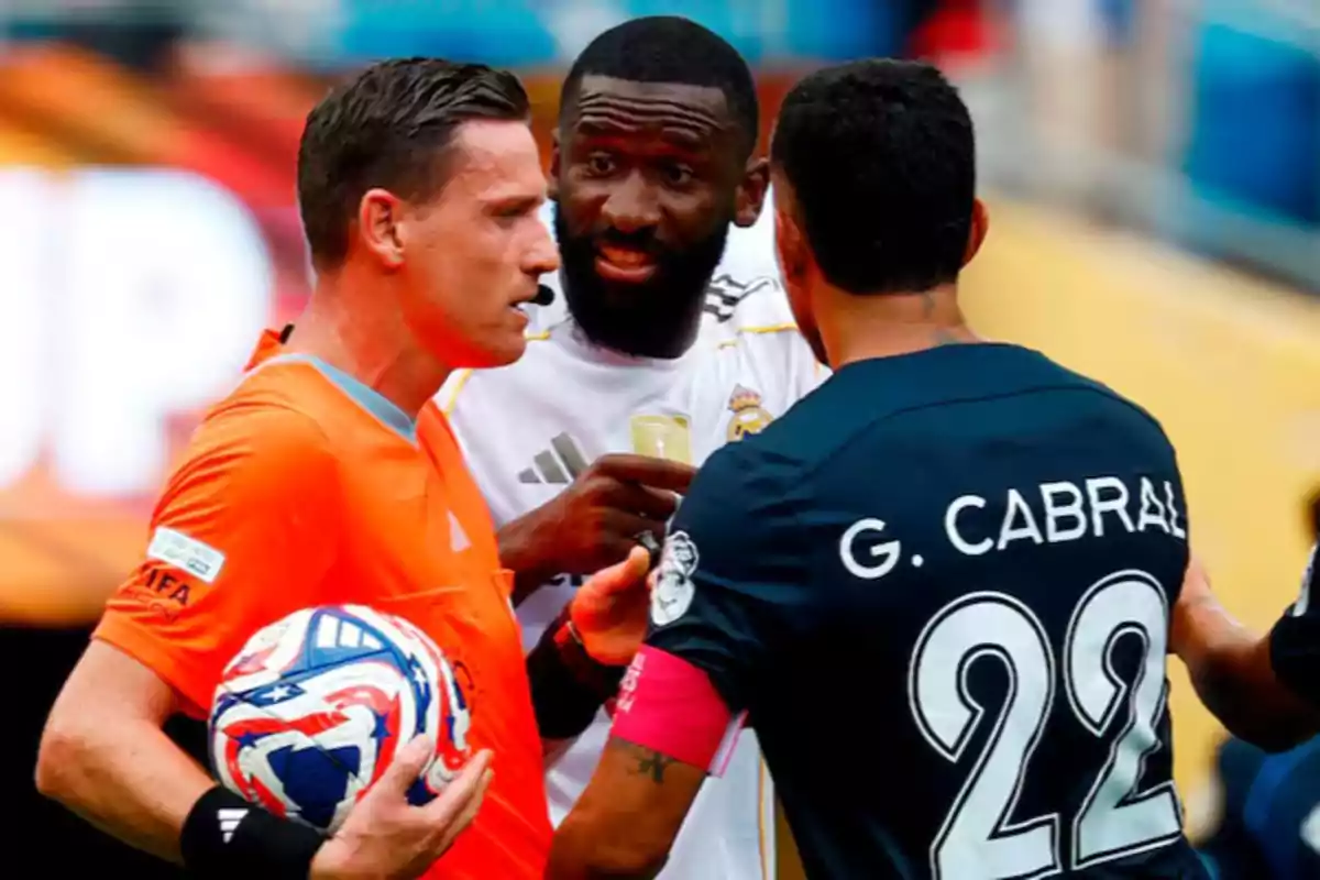 A referee holds a ball while talking with two players during a match.