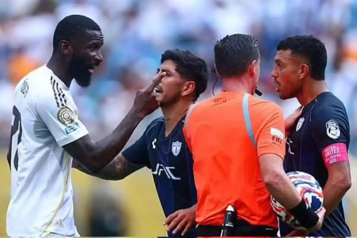 Un jugador de fútbol con uniforme blanco discute acaloradamente señalando el rostro de un rival mientras el árbitro y otro jugador observan la situación en el campo.
