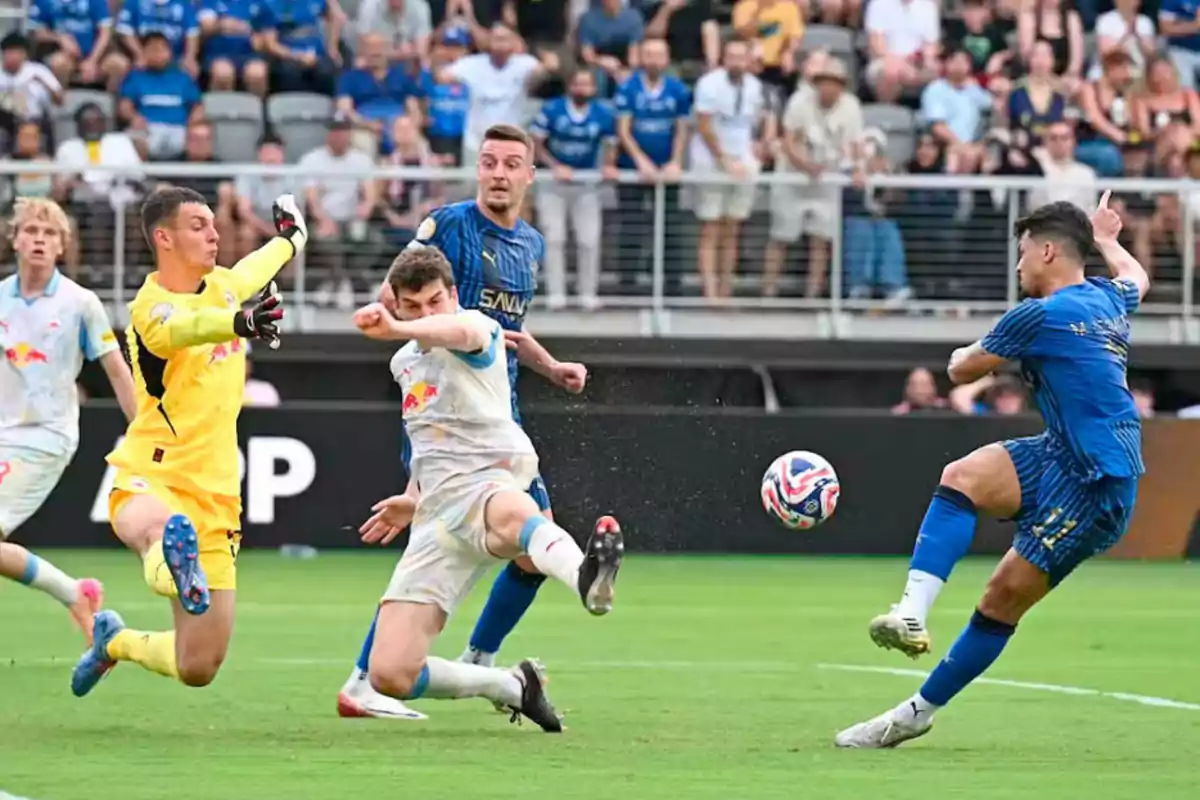 Un jugador de fútbol con uniforme azul dispara el balón hacia la portería mientras el portero y dos defensores con uniforme blanco intentan bloquear el tiro en un estadio lleno de espectadores. Un jugador de fútbol con uniforme azul dispara el balón hacia la portería mientras el portero y dos defensores con uniforme blanco intentan bloquear el tiro en un estadio lleno de espectadores.