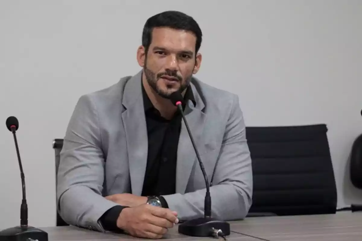 Hombre con barba y saco gris hablando frente a un micrófono en una sala de reuniones Hombre con barba y saco gris hablando frente a un micrófono en una sala de reuniones