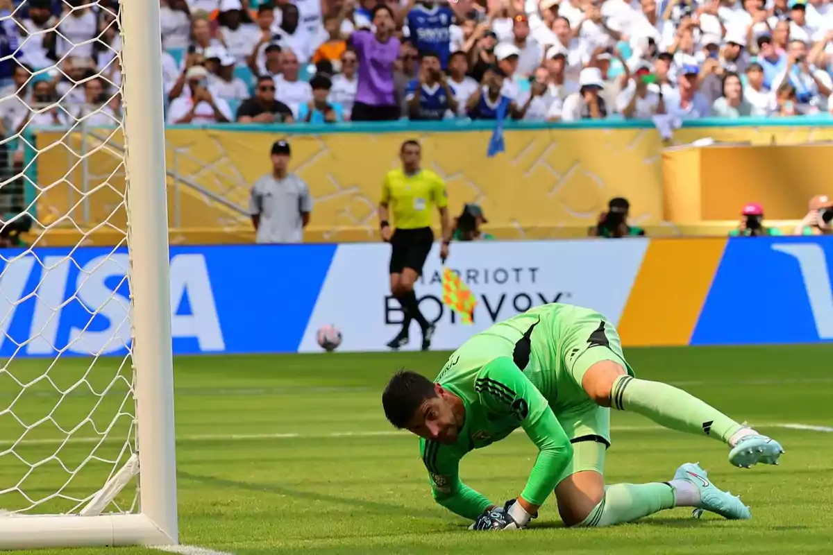 Portero vestido de verde en el césped tras intentar detener un balón durante un partido de fútbol con público y árbitro al fondo Portero vestido de verde en el césped tras intentar detener un balón durante un partido de fútbol con público y árbitro al fondo